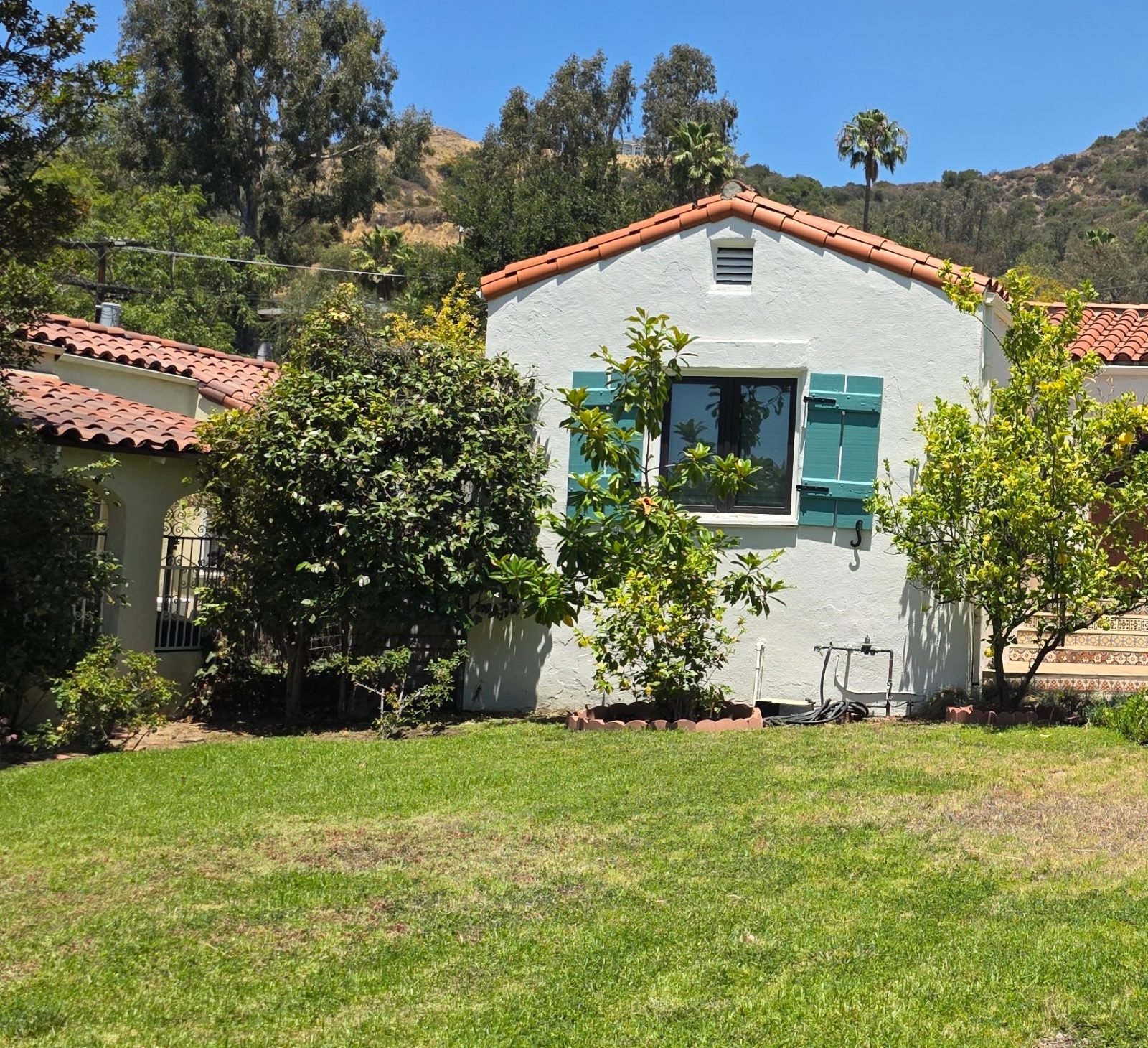 White Spanish style bungalow with teal shutters and a red tile roof behind a front lawn in Eagle Rock, Los Angeles